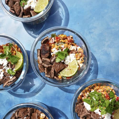 Five glass bowls containing beef strips, roasted corn, crumbled cheese, cilantro, and lime wedges are arranged on a bright blue surface with strong sunlight casting shadows.