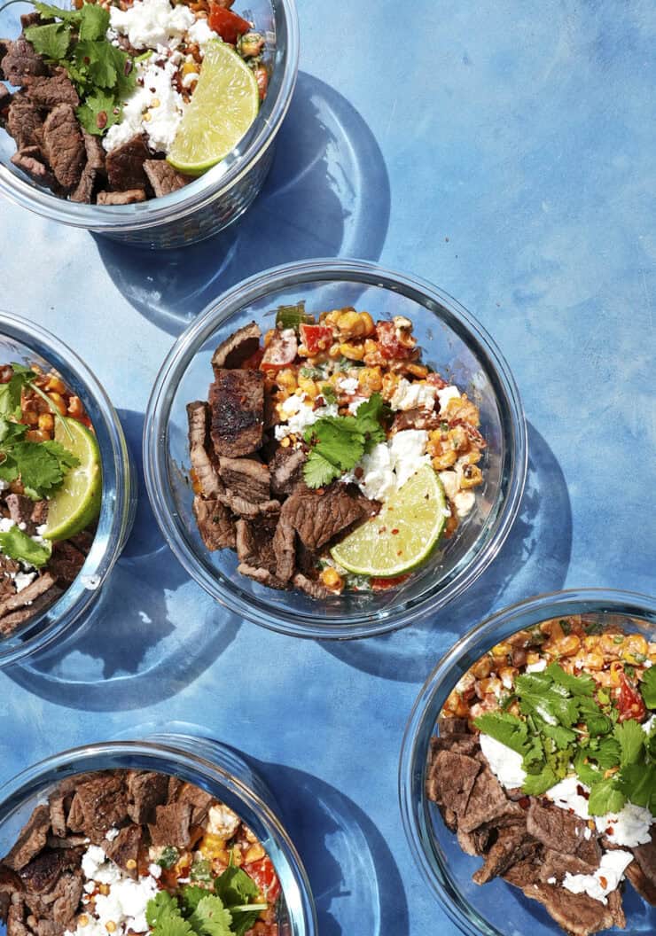 Five glass bowls containing beef strips, roasted corn, crumbled cheese, cilantro, and lime wedges are arranged on a bright blue surface with strong sunlight casting shadows.