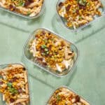 Six glass meal prep containers filled with baked pasta topped with roasted corn, chopped green onions, and bits of bacon or sausage, arranged on a light green surface.