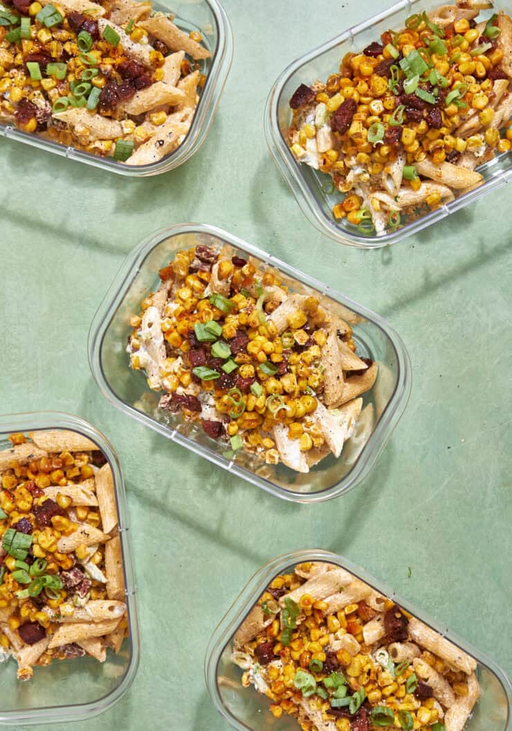 Six glass meal prep containers filled with baked pasta topped with roasted corn, chopped green onions, and bits of bacon or sausage, arranged on a light green surface.