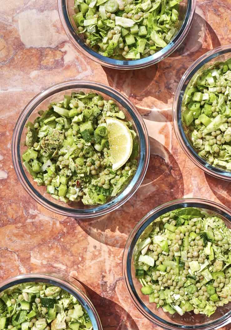 Five glass bowls filled with chopped green vegetables and lentils are arranged on a pink marble surface, with one bowl containing a lemon wedge. Bright sunlight casts distinct shadows.