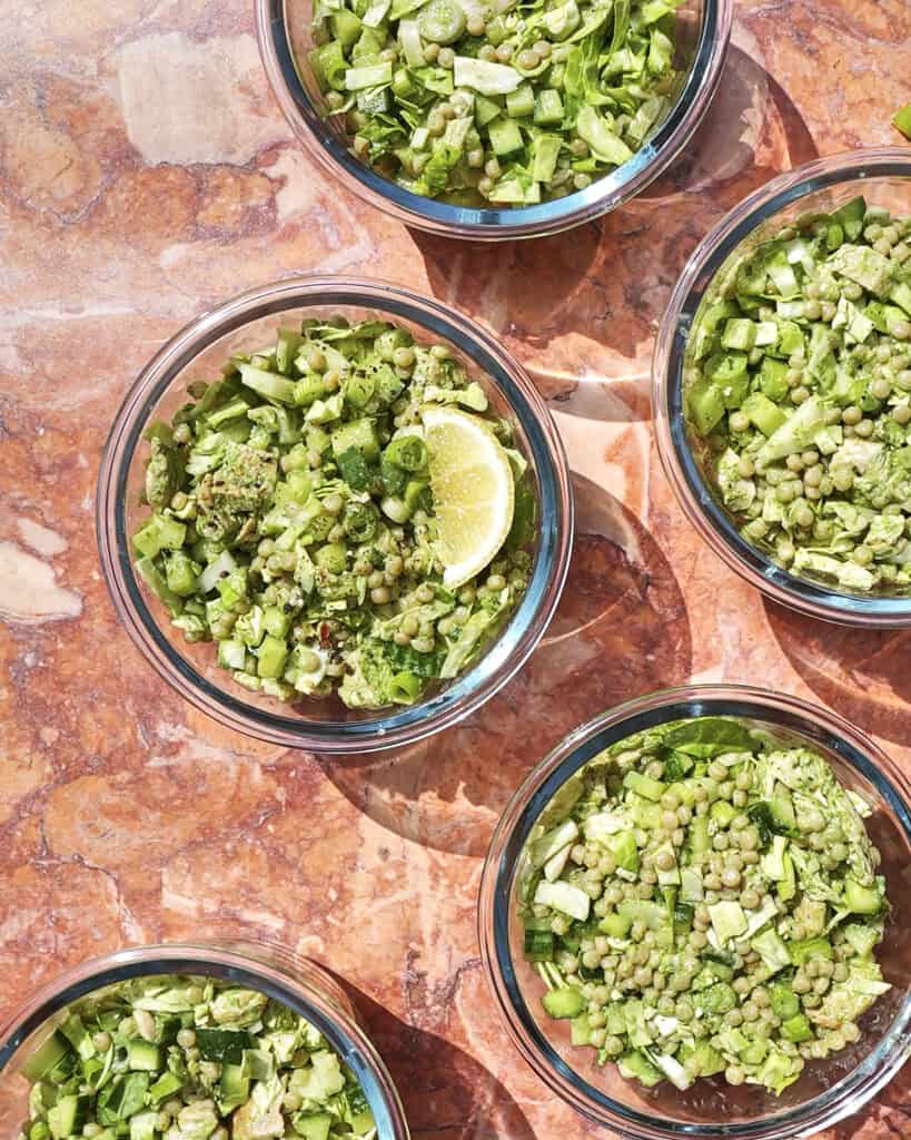 Five glass bowls filled with chopped green vegetables and lentils are arranged on a pink marble surface, with one bowl containing a lemon wedge. Bright sunlight casts distinct shadows.