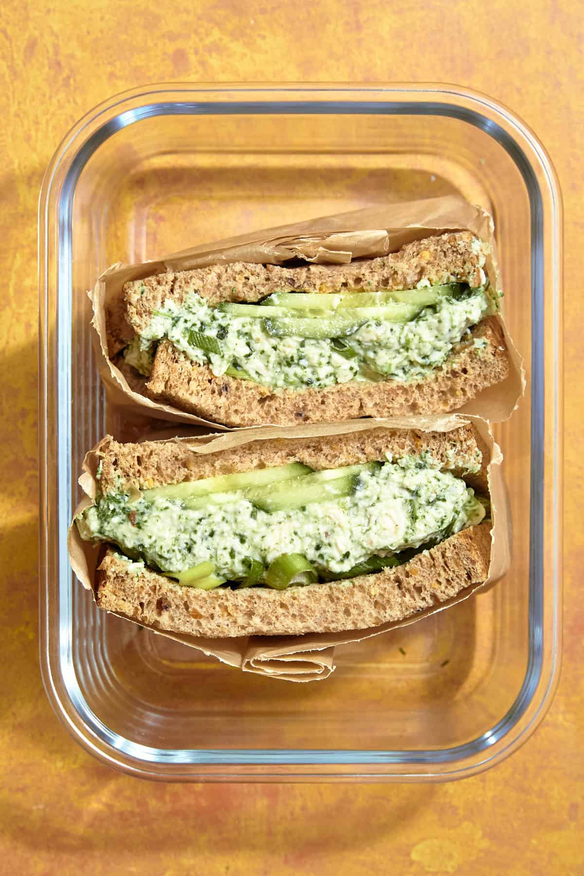 Two halves of a sandwich with green herb spread, roasted corn, and cucumber slices on brown bread, placed in a glass container lined with brown paper, set on an orange surface.