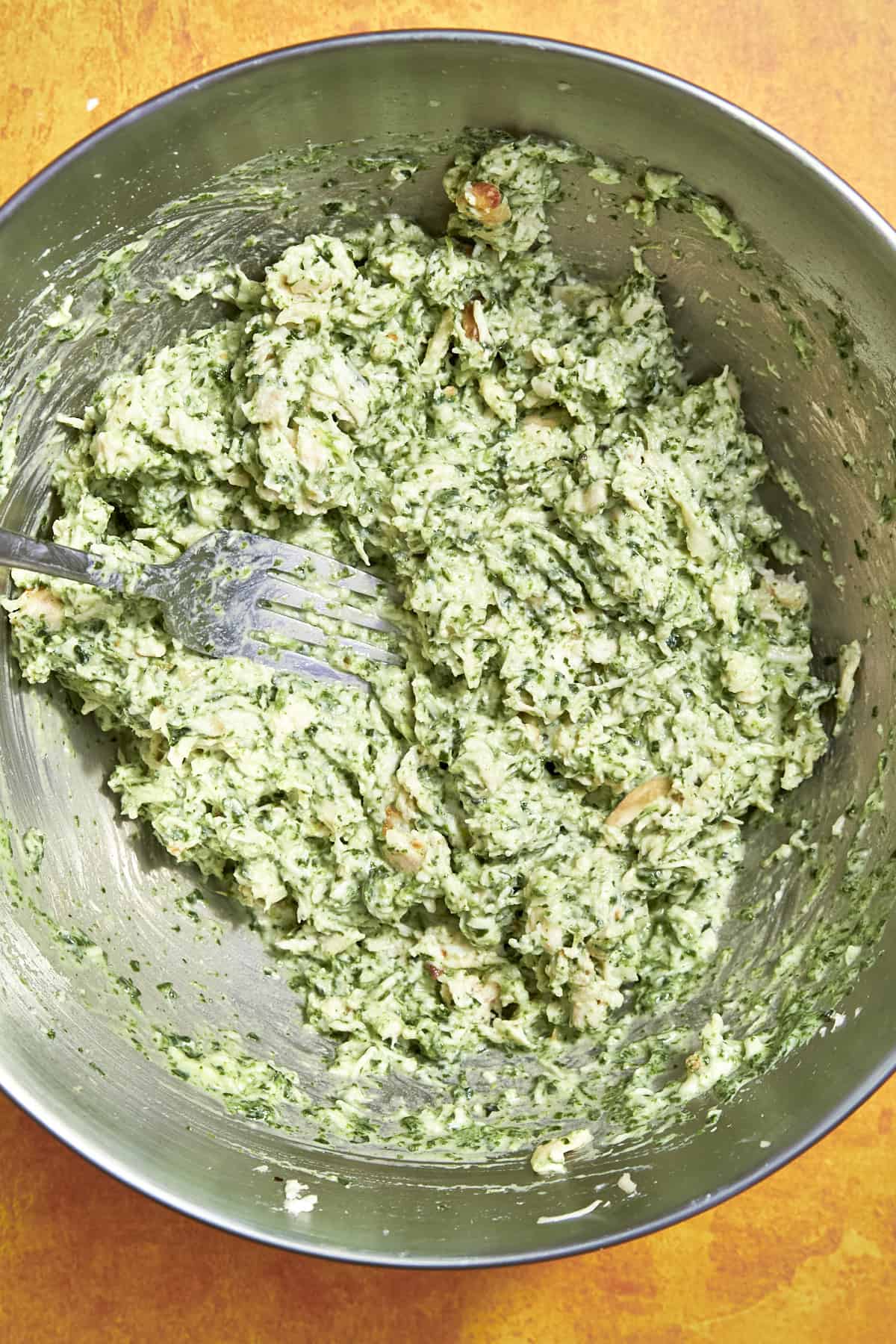 A metal mixing bowl filled with a green and white mixture, likely a spinach, cheese, and roasted corn filling, being stirred with a fork on an orange surface.
