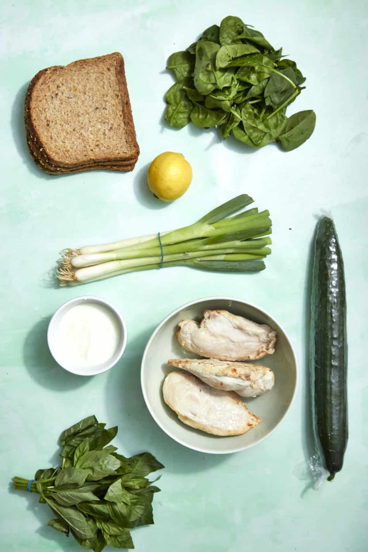 Slices of whole wheat bread, fresh spinach, a lemon, green onions, a cucumber, roasted corn, a bowl of cooked chicken breast, basil, and a small bowl of yogurt are arranged on a light green background.