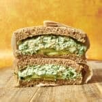 A thick sandwich cut in half, filled with a creamy green mixture, sliced avocado, and roasted corn, sits on a wooden surface against a bright orange background. The sandwich is made with whole grain bread and wrapped in parchment paper.