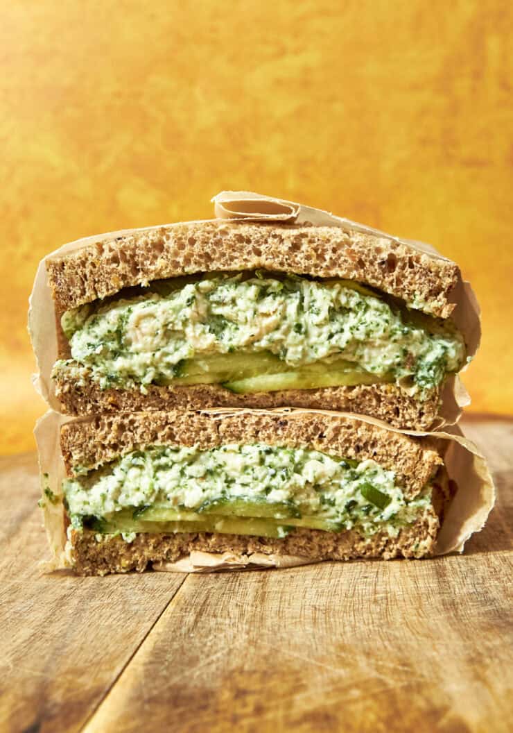 A thick sandwich cut in half, filled with a creamy green mixture, sliced avocado, and roasted corn, sits on a wooden surface against a bright orange background. The sandwich is made with whole grain bread and wrapped in parchment paper.