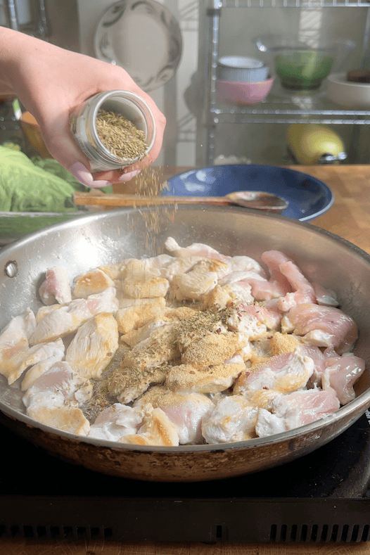 A hand sprinkles dried herbs from a jar onto pieces of raw and partially cooked chicken in a skillet on a stovetop, with kitchen items and fresh lettuce visible in the background.