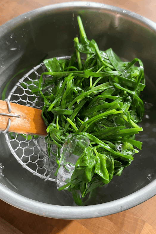 Blanched leafy greens rest on a slotted ladle over a metal bowl filled with ice water, set on a wooden surface.