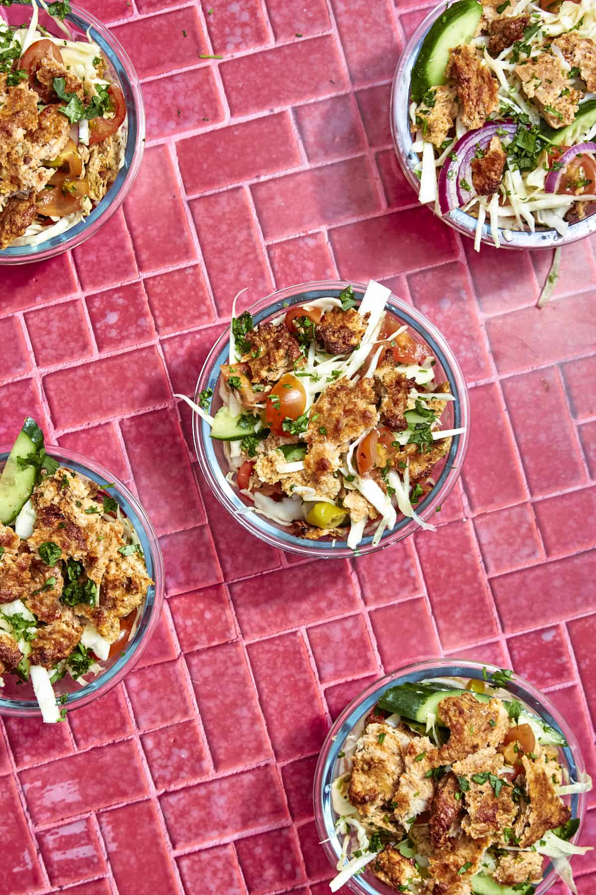 Five glass bowls filled with fresh salad, including tomatoes, cucumbers, cabbage, greens, crispy pieces, and roasted corn, are arranged on a bright pink tiled surface.