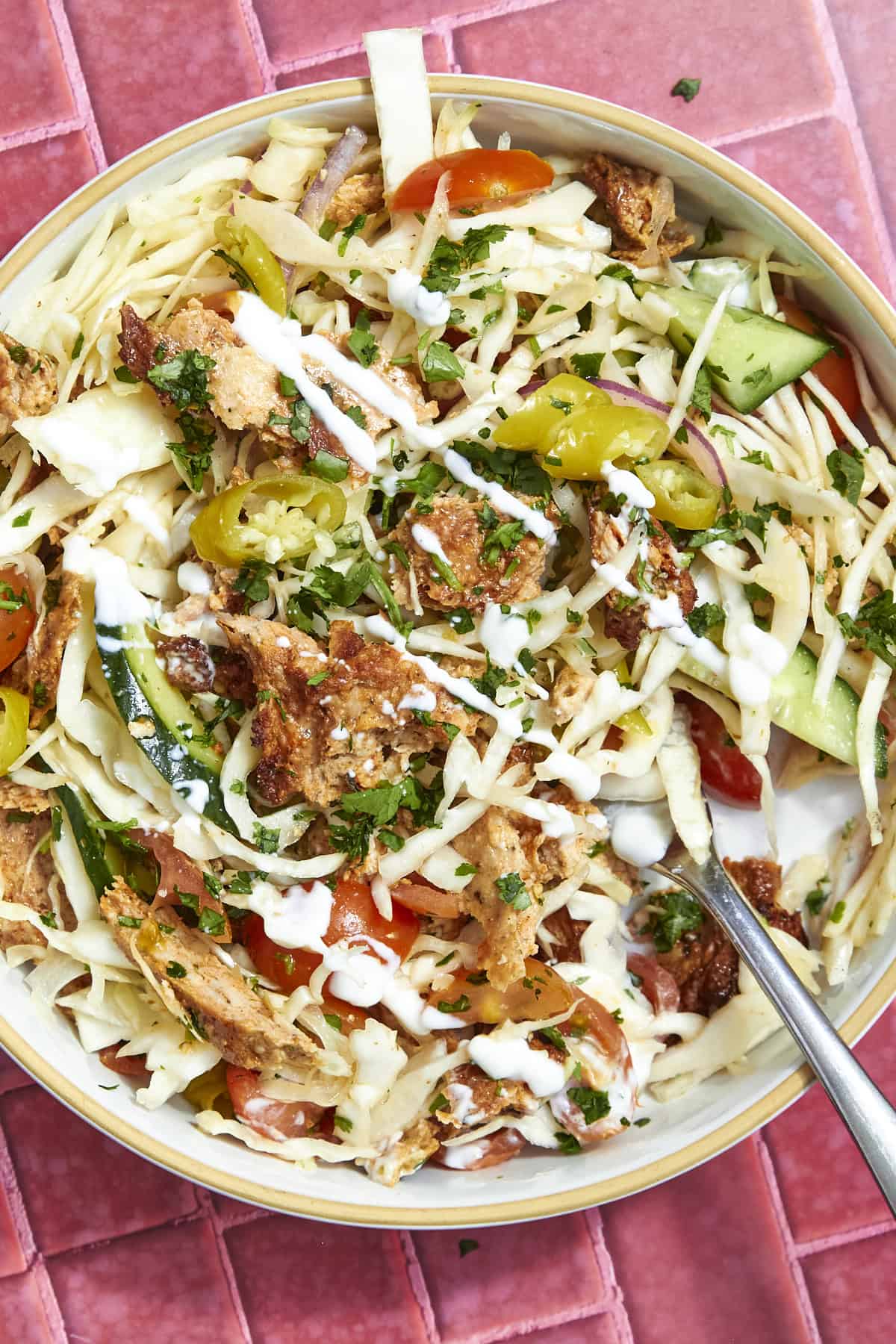 A bowl of chopped salad with cabbage, cucumbers, tomatoes, pepperoncini, roasted corn, crispy pita chips, fresh herbs, and a drizzle of creamy dressing, set on a pink tiled surface with a spoon in the bowl.