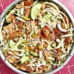 A colorful salad in a metal bowl with shredded cabbage, sliced cucumbers, roasted corn, red onions, cherry tomatoes, jalapeños, herbs, and crispy bread pieces on a red tiled surface.