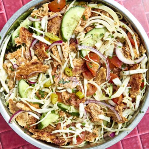 A colorful salad in a metal bowl with shredded cabbage, sliced cucumbers, roasted corn, red onions, cherry tomatoes, jalapeños, herbs, and crispy bread pieces on a red tiled surface.