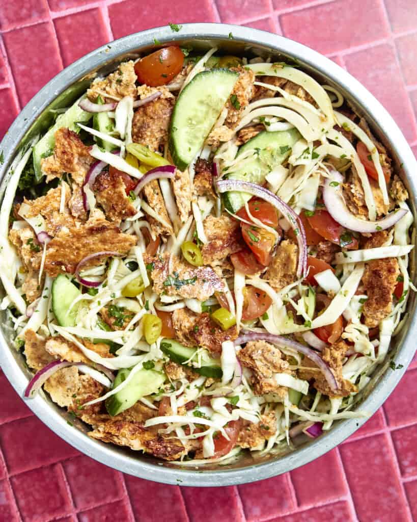 A colorful salad in a metal bowl with shredded cabbage, sliced cucumbers, roasted corn, red onions, cherry tomatoes, jalapeños, herbs, and crispy bread pieces on a red tiled surface.