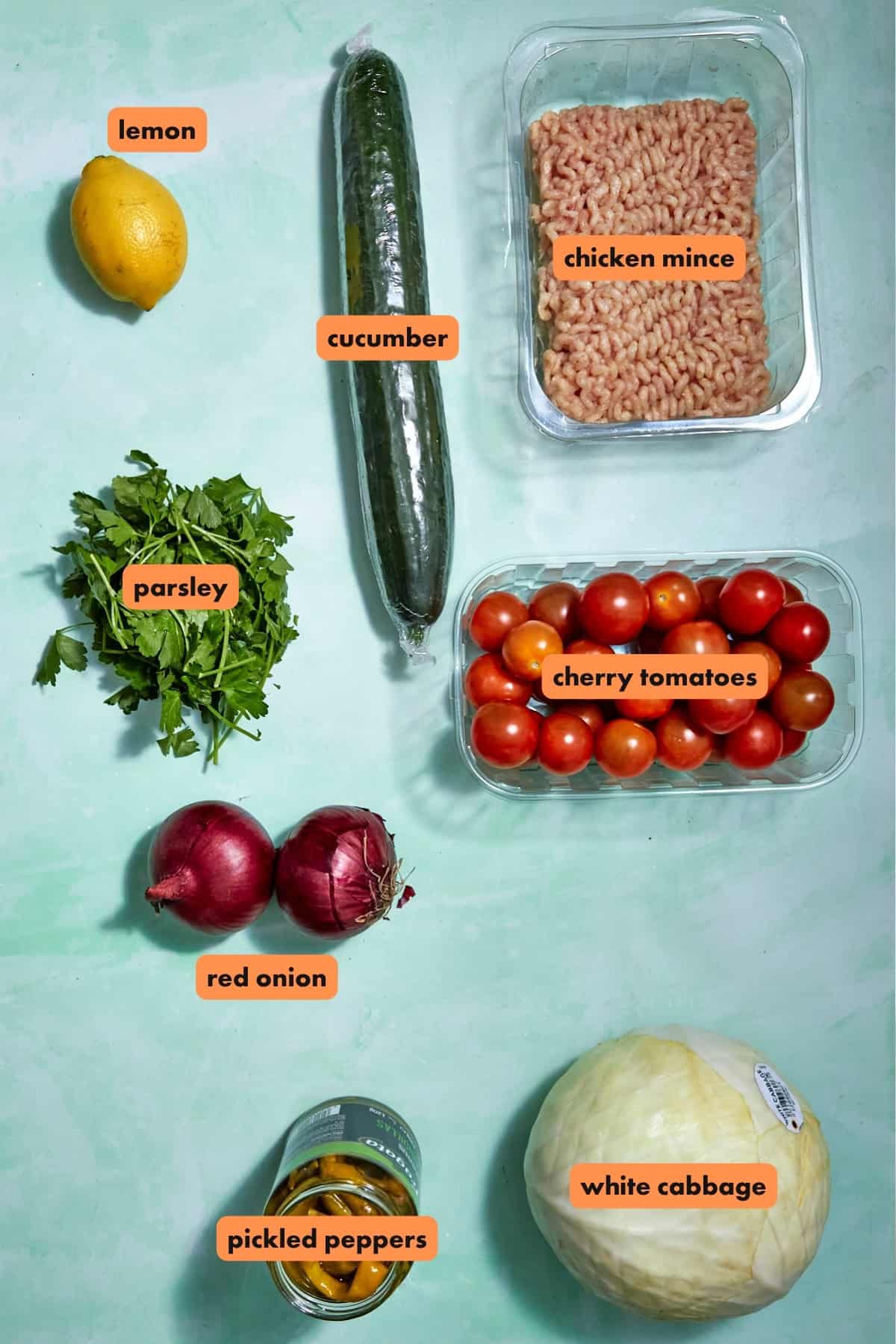 A lemon, cucumber, tray of chicken mince, bunch of parsley, container of cherry tomatoes, two red onions, jar of pickled peppers, roasted corn, and a head of white cabbage are arranged on a light green surface with labels.
