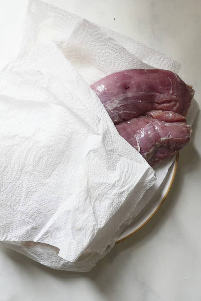 Raw pork tenderloin partially covered with paper towels on a white plate, set on a light-colored countertop, ready to be seasoned and cooked as delicious pork tenderloin in oven.