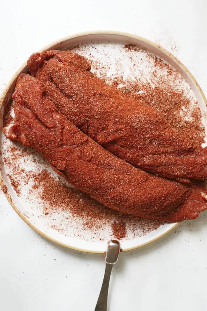 Two raw pork tenderloins, perfect for making pork tenderloin in oven, are coated with a reddish spice rub on a round white plate, extra seasoning scattered around and a metal serving fork resting alongside.