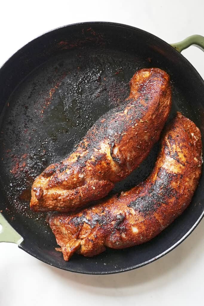 Two seared pork tenderloins coated in spices cook in a cast iron skillet, showing a dark, crusty exterior with char marks—perfect for finishing as pork tenderloin in oven. The pan has a green handle and sits on a white background.