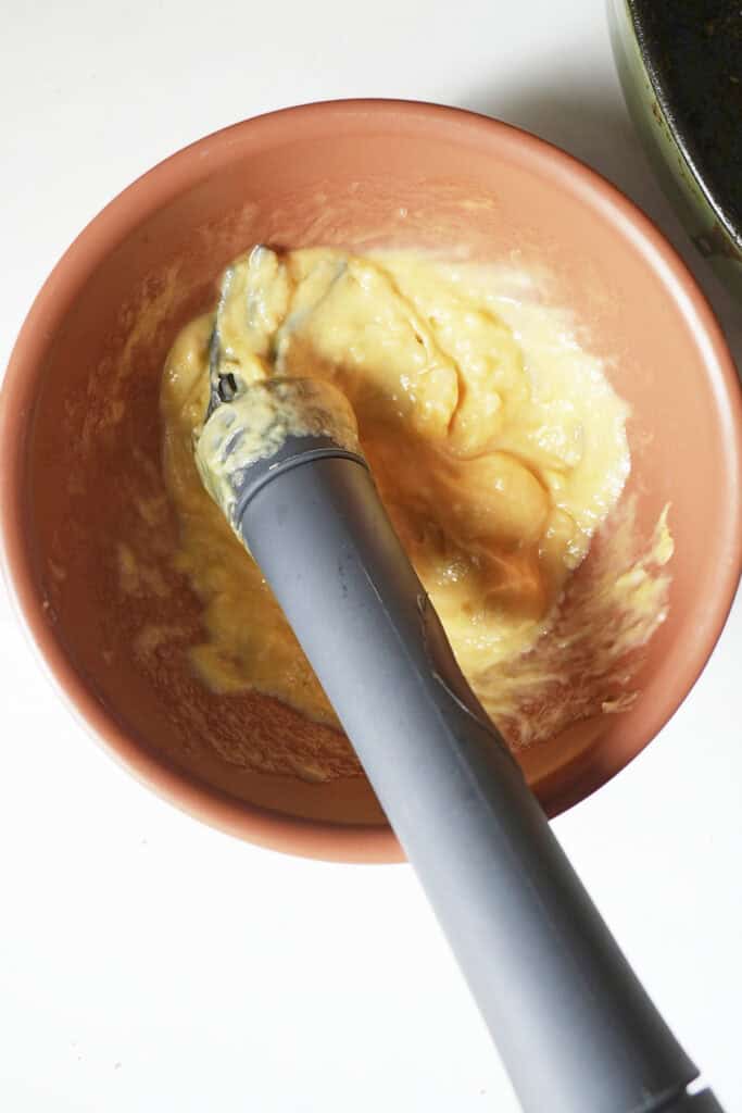 A silicone spatula is mixing eggs in a terracotta-colored bowl beside a tray of pork tenderloin in oven. The eggs are partially beaten, with visible streaks of yellow yolk and white. The bowl rests on a white surface.