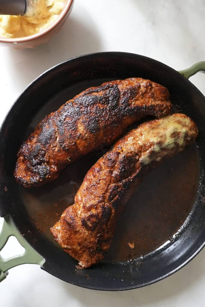 Two browned pork tenderloins with a crispy crust are cooking in a black cast iron pan, fresh from finishing as pork tenderloin in oven. One has a melted, golden area on top. The pan rests on a white surface, with a bowl visible in the corner.