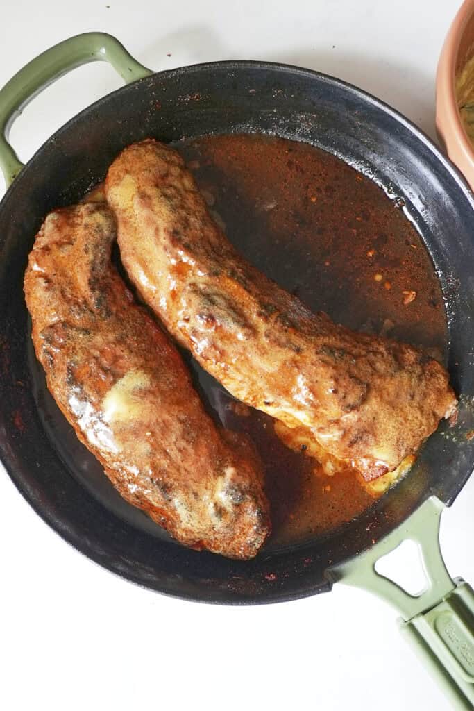 Two cooked, sauced pork tenderloins in a round black skillet with green handles, fresh from the oven, sit in a pool of reddish-brown sauce on a white surface.