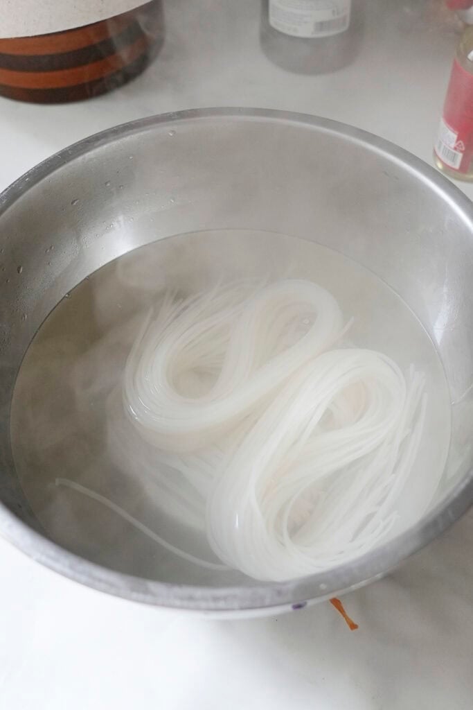 A metal bowl filled with hot water and soaking white rice noodles sits on the kitchen counter, steam rising gently while a hint of roasted corn aroma lingers in the air.