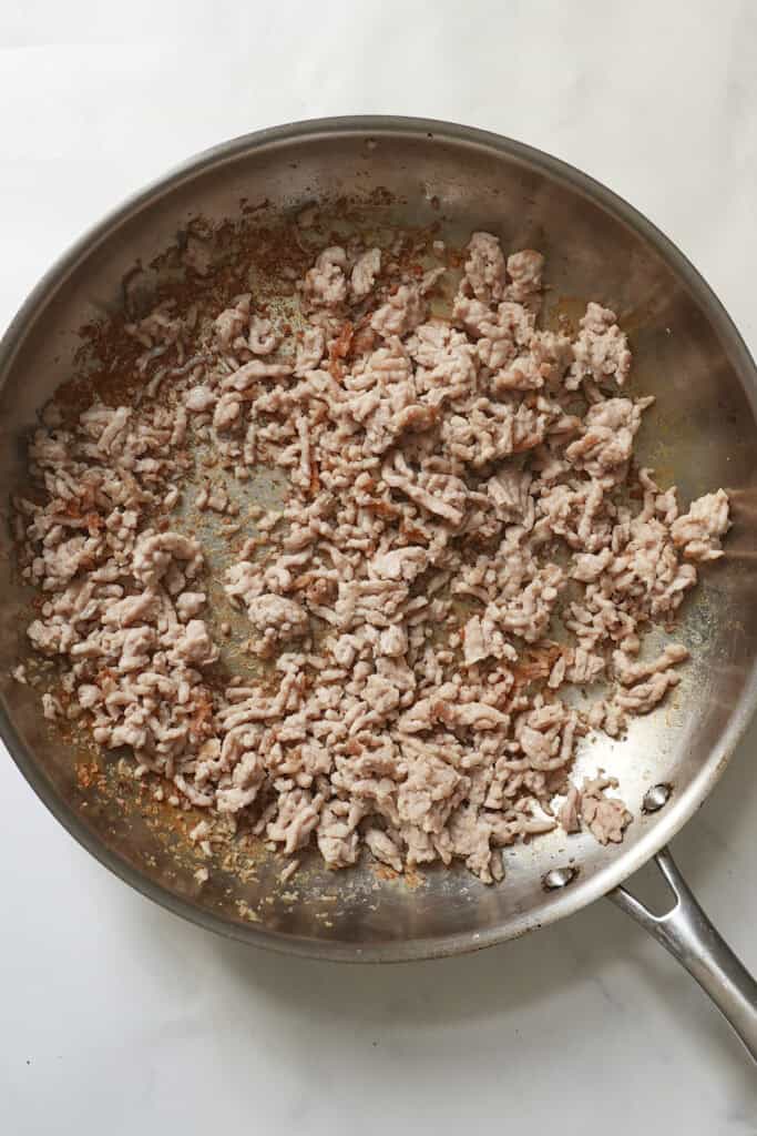 Ground meat browning in a stainless steel skillet on a white surface, with some browned bits, juices, and scattered roasted corn adding extra color and flavor to the pan.