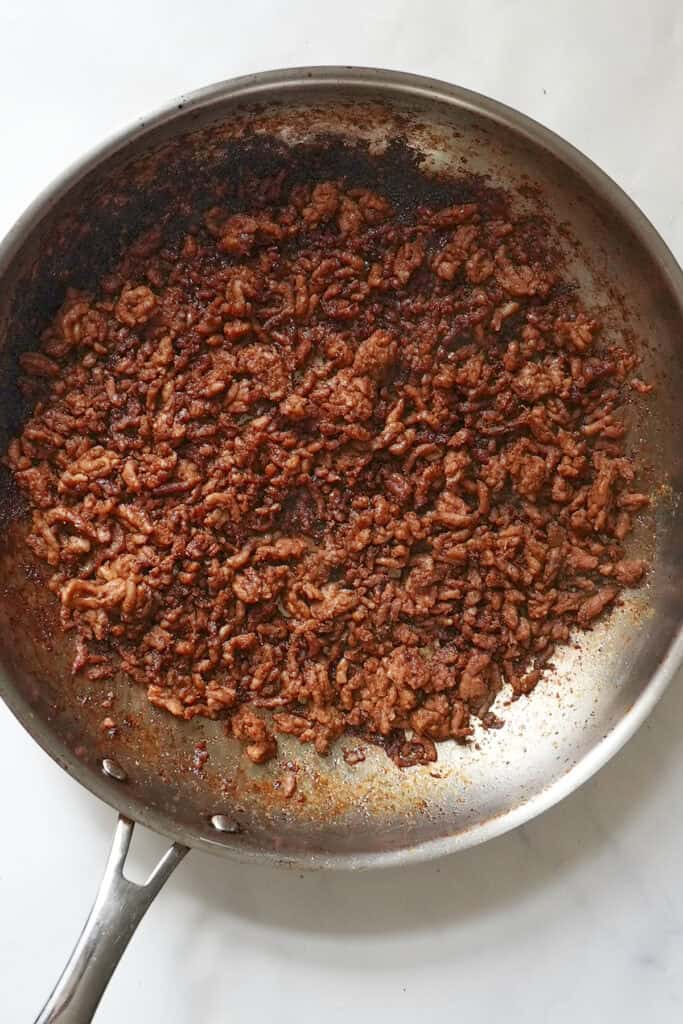 Cooked and browned ground beef with spices and roasted corn in a stainless steel skillet, resting on a white surface.