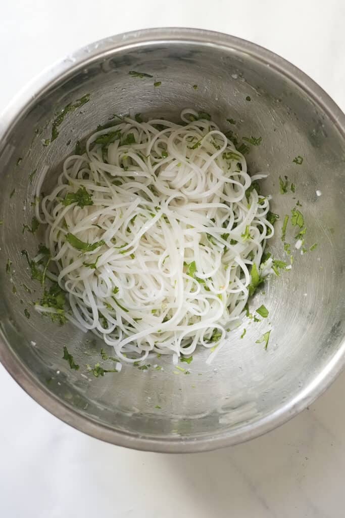 A metal bowl filled with cooked rice noodles, roasted corn, and chopped fresh herbs, placed on a light-colored surface.