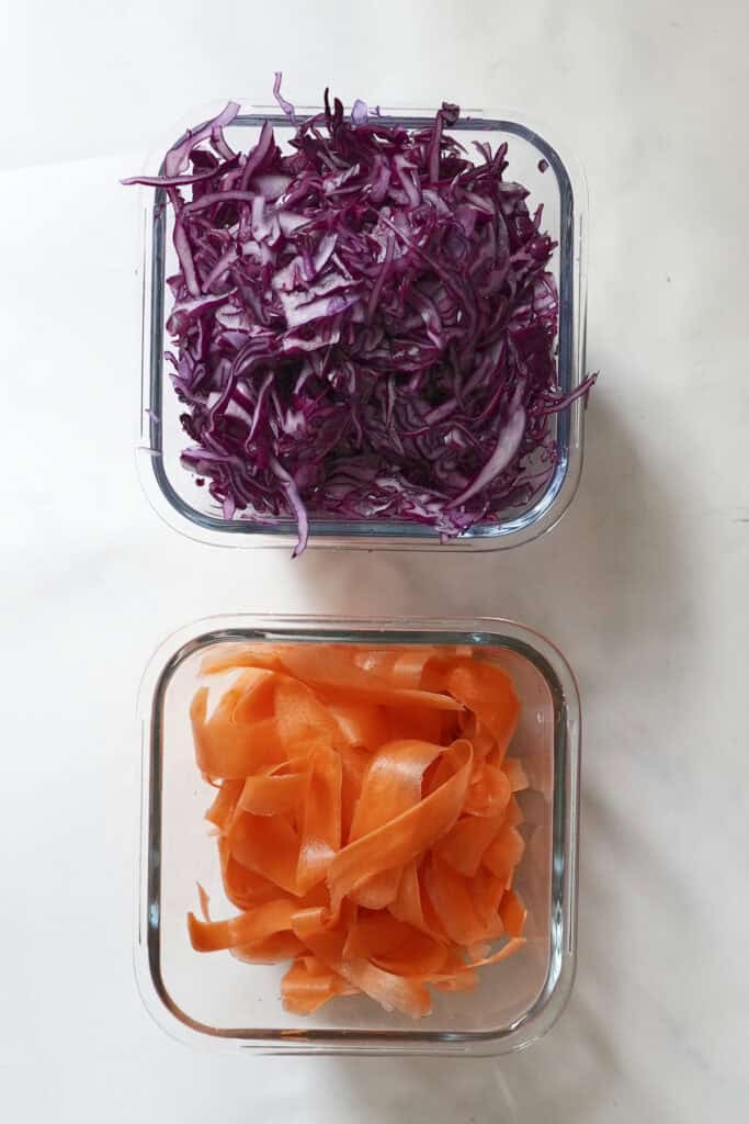 Two glass containers on a white surface: the top holds shredded purple cabbage, while the bottom holds wide, ribbon-like carrot slices beside a sprinkle of roasted corn for added texture and flavor.