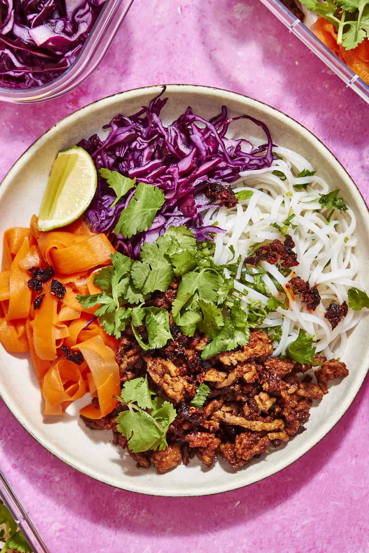A bowl with rice noodles, roasted corn, shredded purple cabbage, carrot ribbons, seasoned crumbled tofu, fresh cilantro, a lime wedge, and chili oil, arranged on a pink surface.