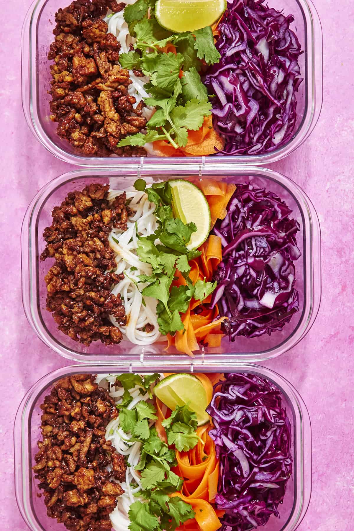 Three meal prep containers filled with cooked ground meat, rice noodles, roasted corn, shredded purple cabbage, carrot ribbons, fresh cilantro, and lime wedges, arranged neatly on a bright pink background.