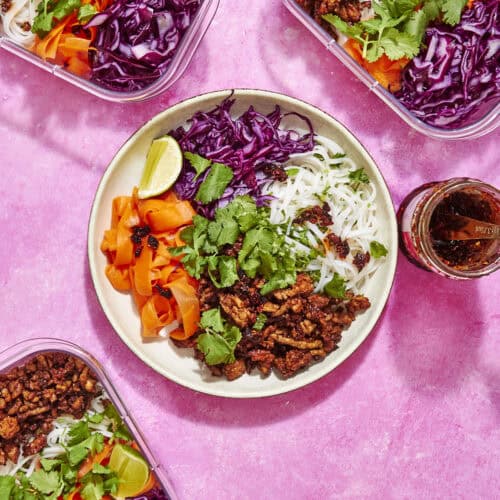 A bowl and three containers filled with rice noodles, roasted corn, shredded purple cabbage, carrot ribbons, seasoned ground meat, fresh cilantro, and lime wedges are arranged on a vibrant pink surface next to a jar of chili sauce.