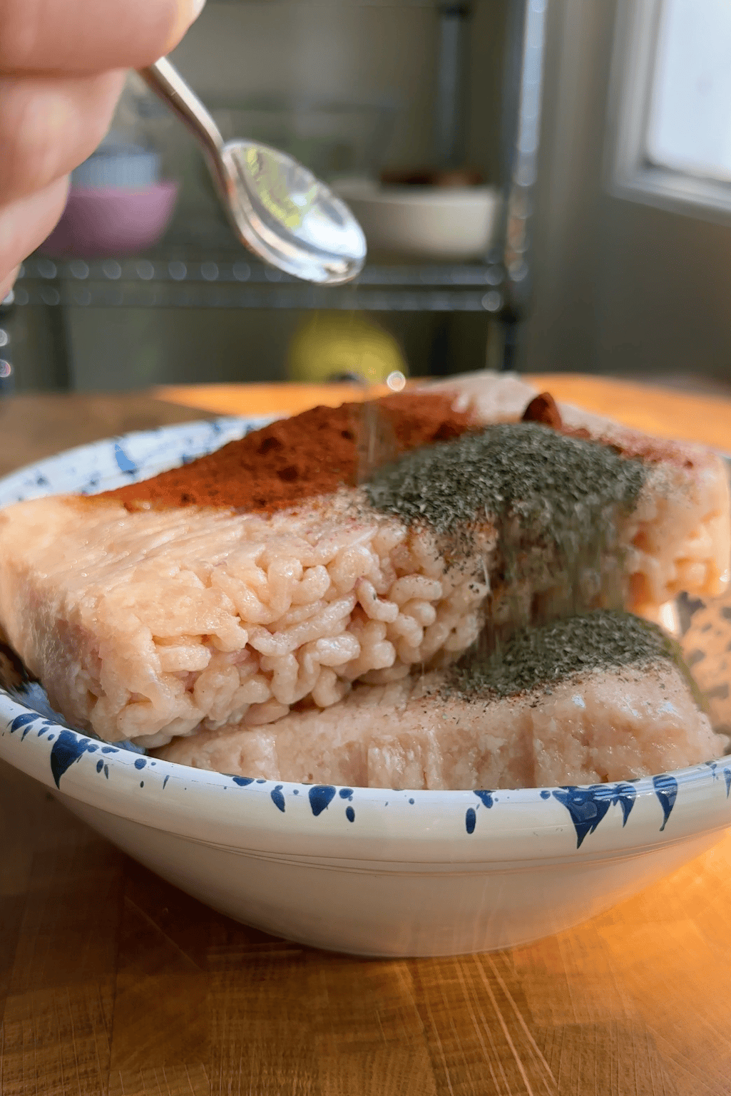 A close-up of a bowl containing pinkish rice blocks topped with green and red powdered spices and bits of roasted corn, with a hand holding a small spoon above the bowl as sunlight shines on the dish.