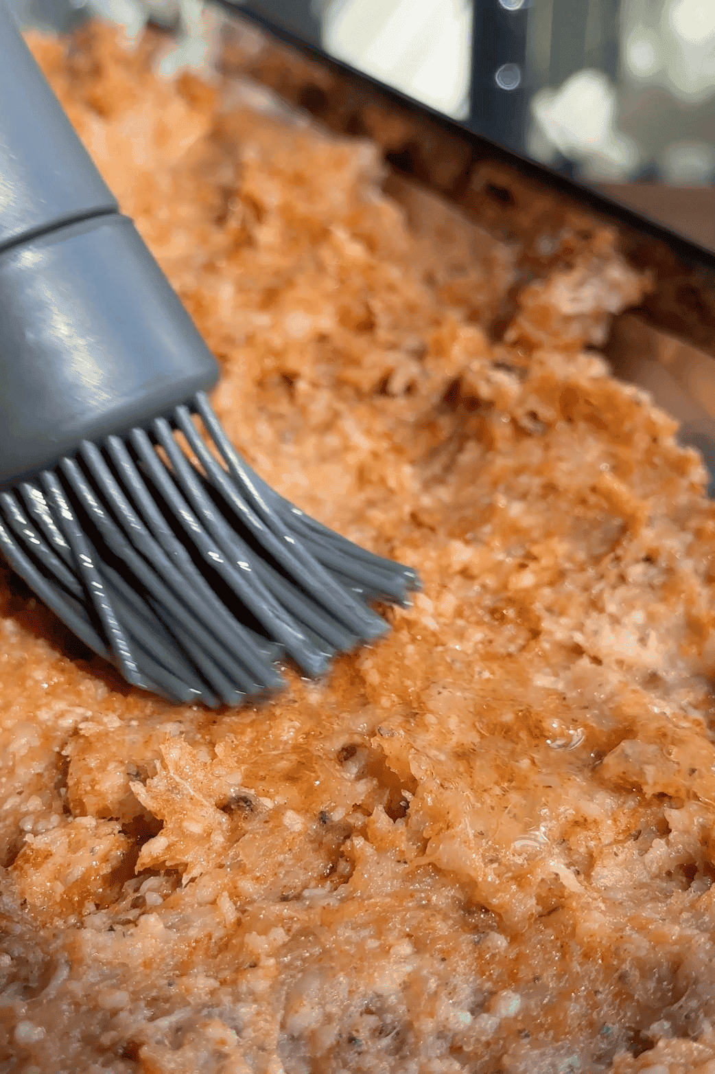 A close-up of a silicone basting brush spreading sauce or oil over a baked, crumbly, golden-brown food surface, possibly a casserole or meatloaf, alongside tender roasted corn for added flavor and texture.