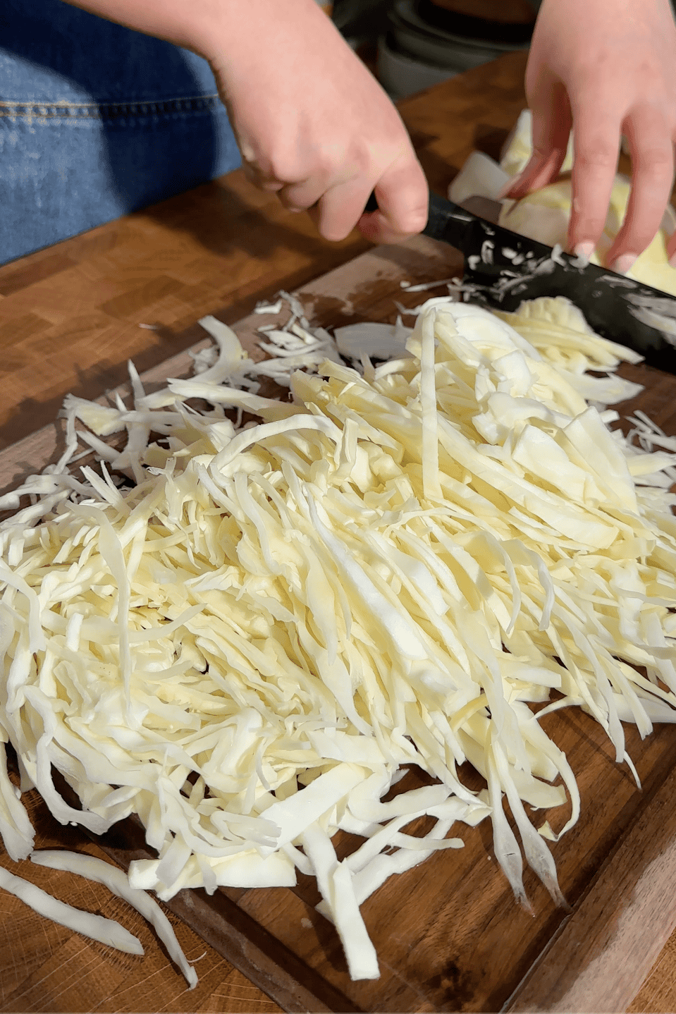 A person uses a large knife to slice fresh cabbage into thin strips on a wooden cutting board, with roasted corn scattered among the shredded cabbage piled in the foreground.