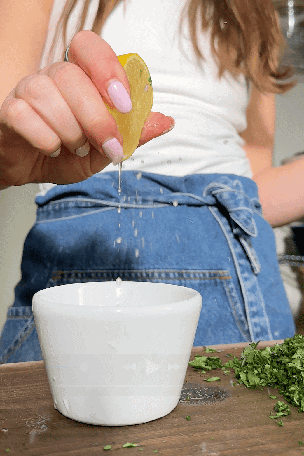 A person squeezes a lemon wedge over a white bowl, with freshly chopped herbs and roasted corn on a wooden surface while wearing a denim apron.