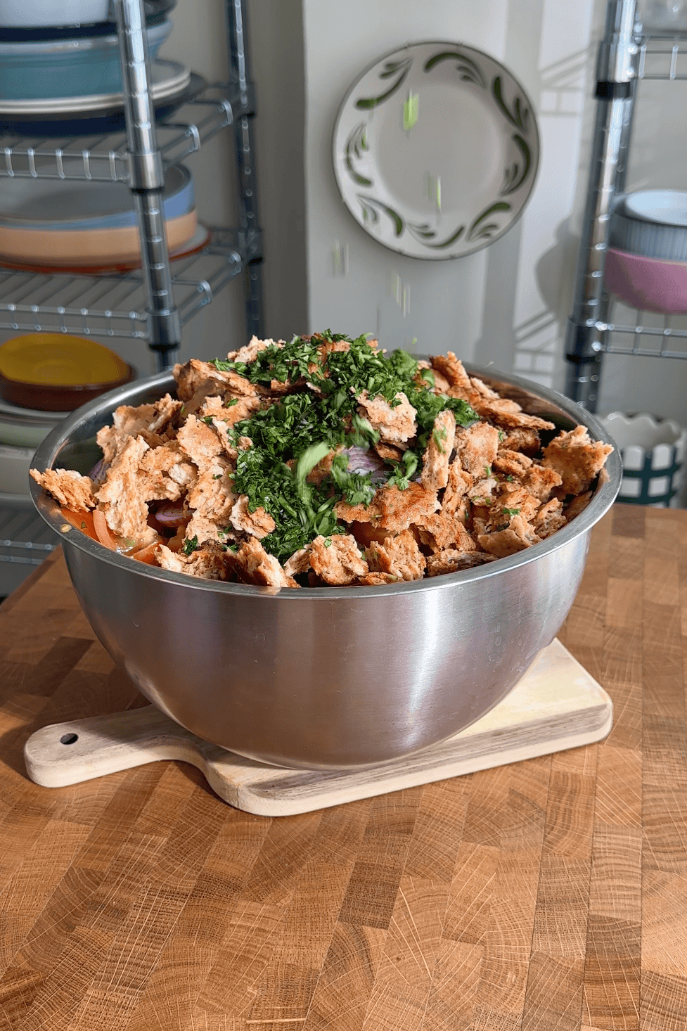 A large metal bowl on a wooden cutting board is filled with torn bread pieces, roasted corn, chopped herbs, and vegetables, set in a kitchen with shelving and dishes in the background.