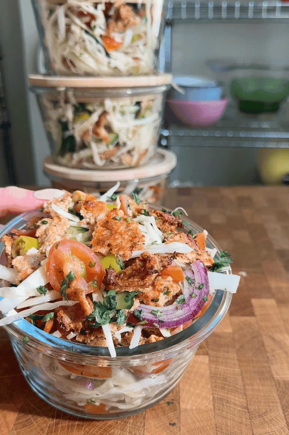 A hand holds a glass bowl filled with a colorful salad featuring roasted corn, shredded cabbage, sliced tomatoes, red onions, herbs, and a crumbled topping. More salad bowls are stacked in the background on a wooden surface.