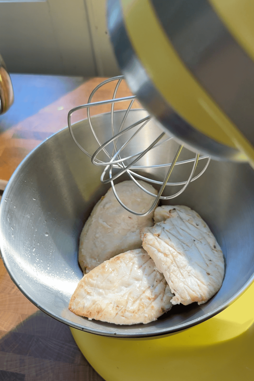 Three cooked chicken breasts sit in the mixing bowl of a stand mixer with a metal whisk attachment, ready for shredding. Nearby, roasted corn adds color as sunlight illuminates the scene, casting slight shadows on the wooden surface below.