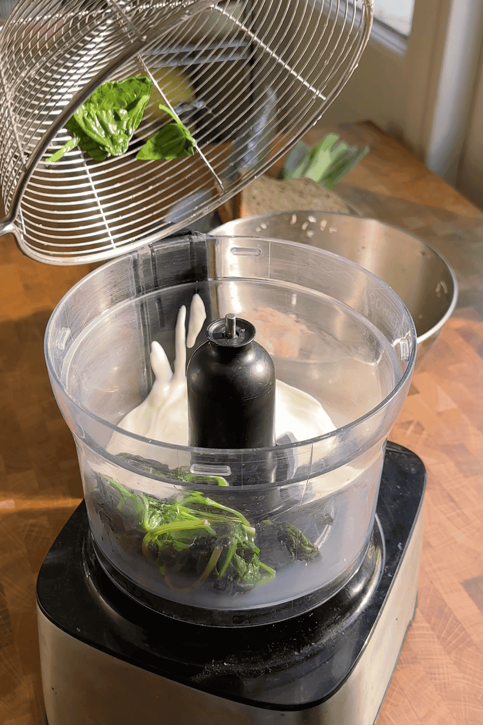 A food processor bowl with green herbs, roasted corn, and a white creamy ingredient sits on a wooden surface. A strainer above is pouring more green herbs into the processor as sunlight shines on the scene.