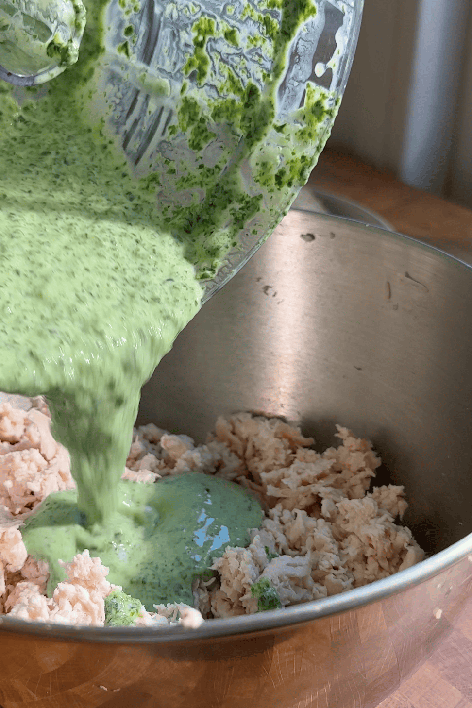 A green herb sauce is being poured from a blender into a metal bowl containing shredded cooked chicken and roasted corn.
