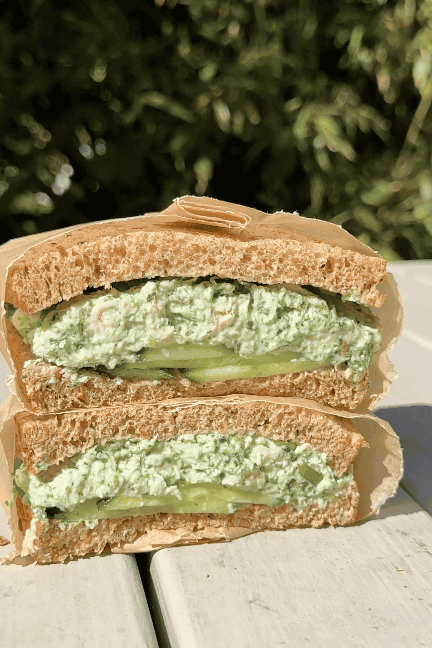 A close-up of a sandwich cut in half, filled with a green herbed spread, cucumber slices, and roasted corn on whole wheat bread. The sandwich is wrapped in brown paper and placed on a white outdoor table with greenery in the background.