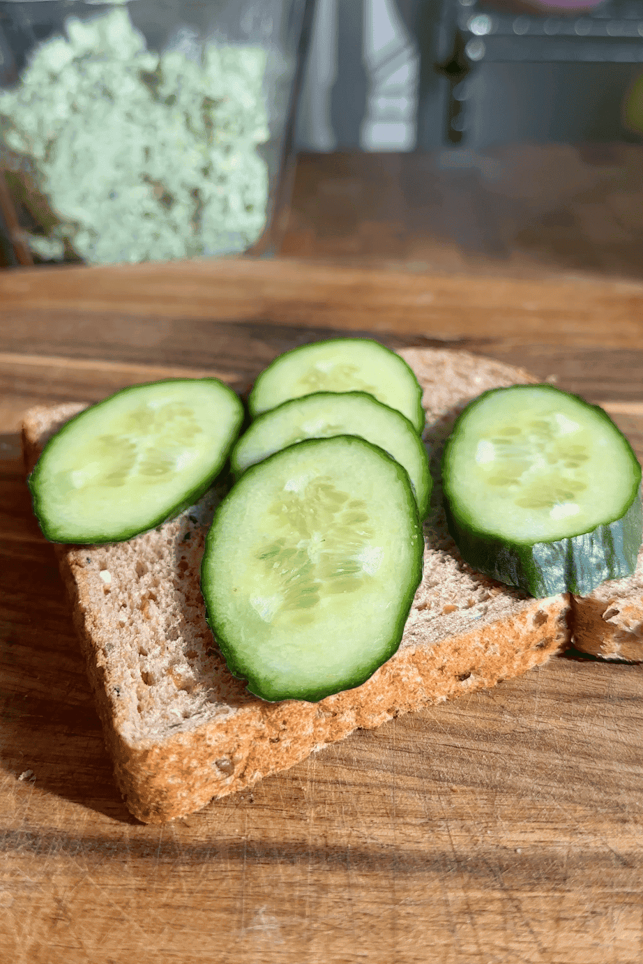 A slice of whole grain bread topped with five slices of fresh cucumber and a hint of roasted corn, placed on a wooden surface. A container with a green spread is blurred in the background.