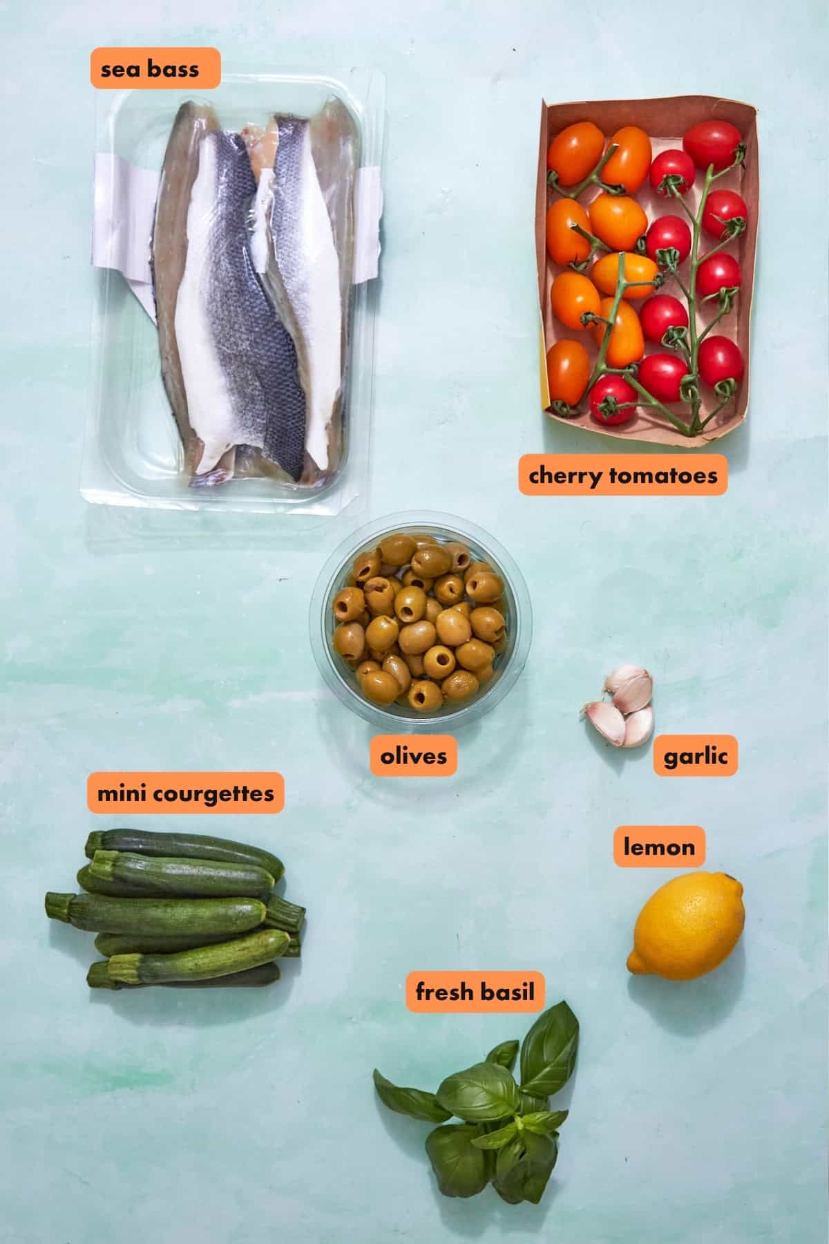 Labeled ingredients on a light surface for a sea bass recipe: raw sea bass fillets, cherry tomatoes on the vine, green olives in a bowl, garlic cloves, a lemon, fresh basil leaves, and several mini courgettes.