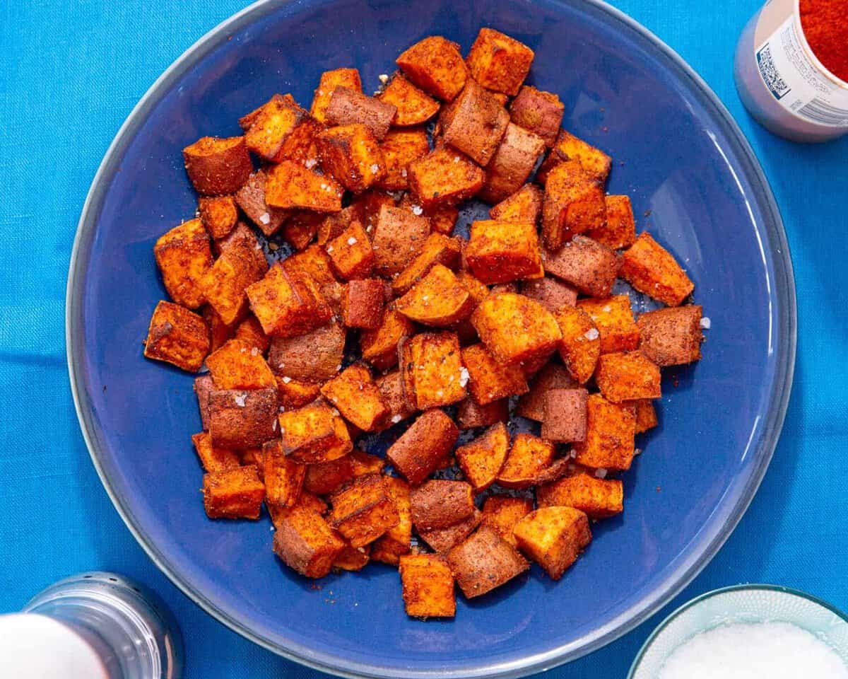 Cooked and browned sweet potato cubes on a blue plate on a blue background next to some salt in a bowl and a container with spices.