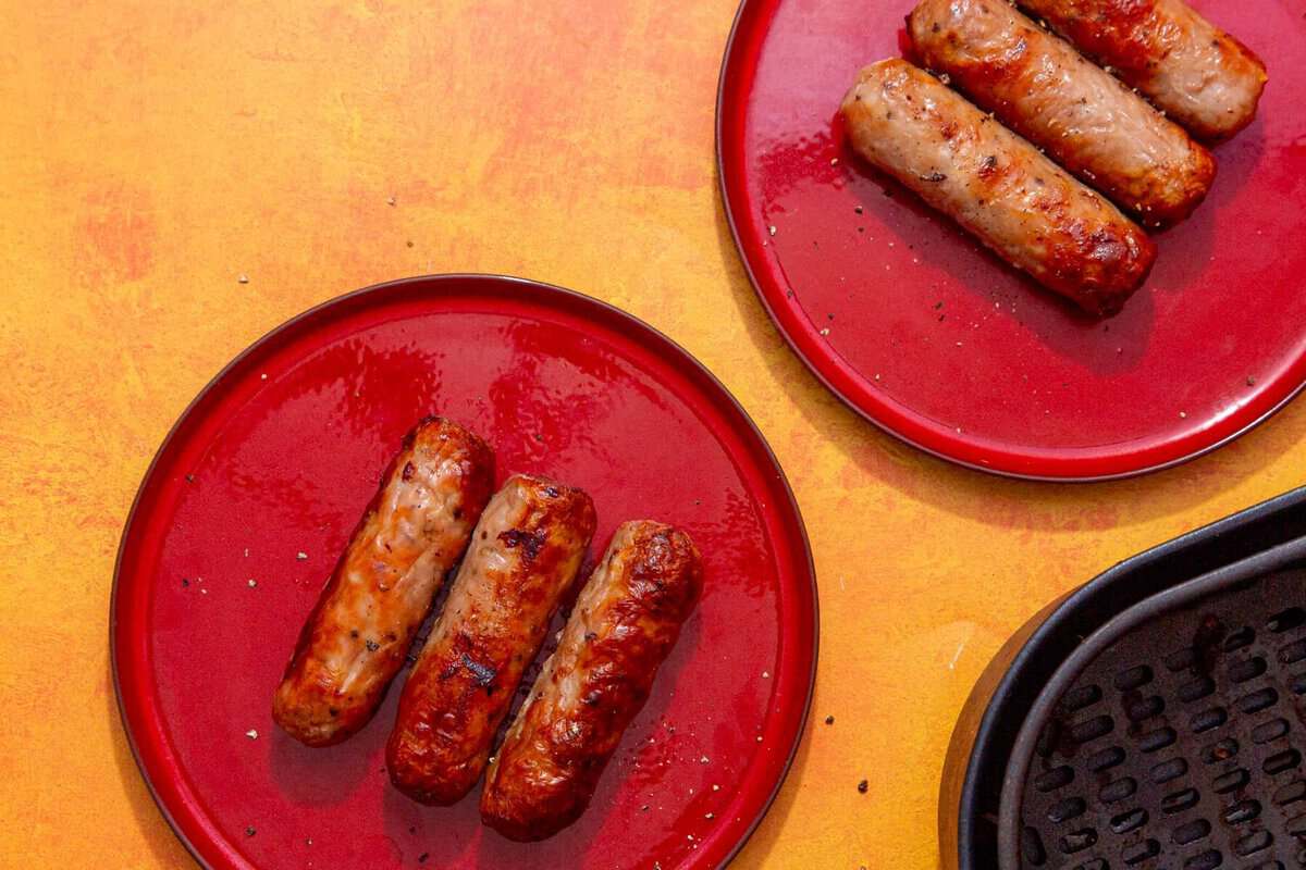 Two red plates, each holding three cooked sausages, are placed on a yellow-orange surface. Part of an air fryer basket appears in the bottom right corner. The sausages look browned and grilled.