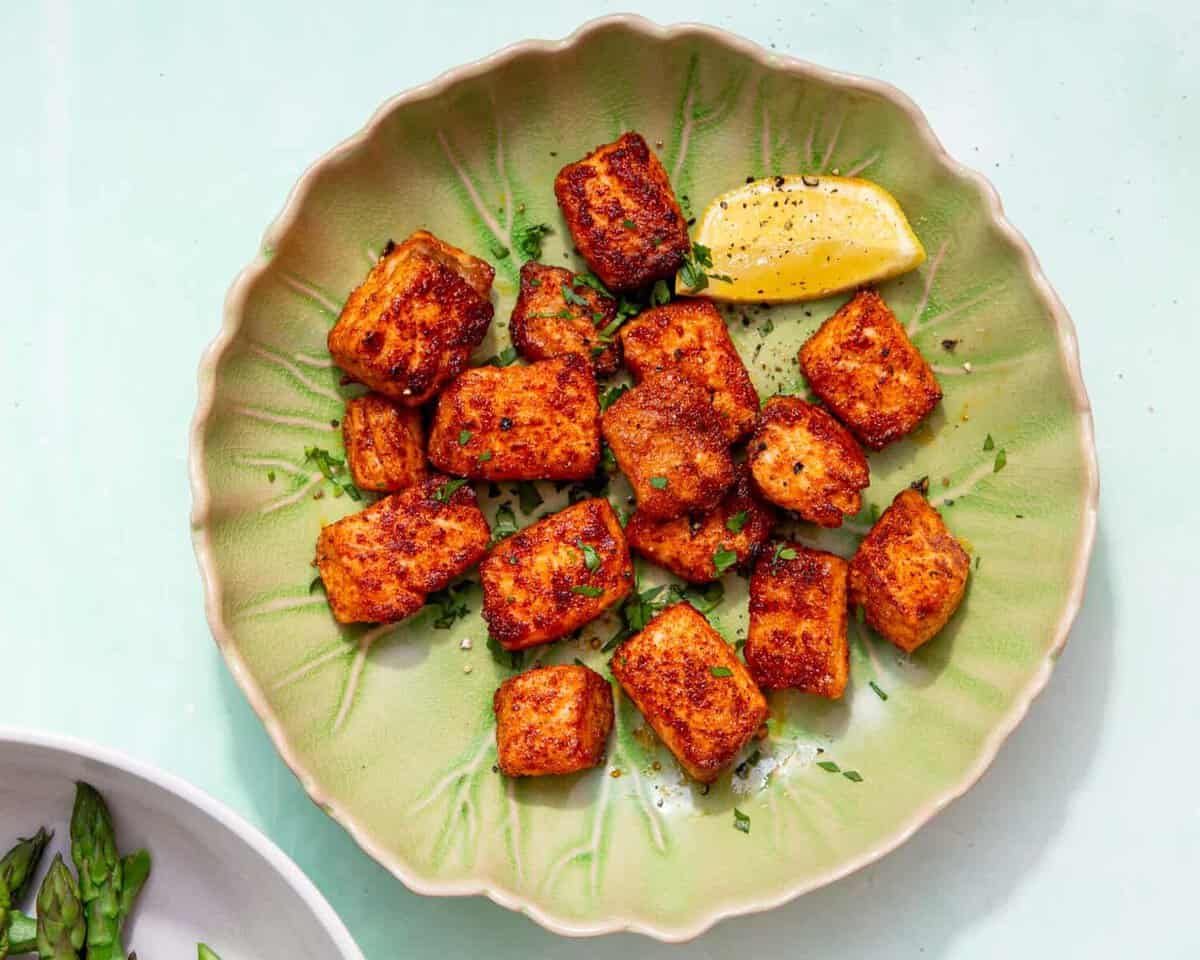 Pieces of golden browned seasoned salmon bites in a green dish with a wedge of lemon.