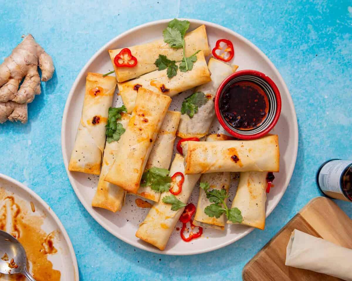 plate of air fryer spring rolls and a cup of soy sauce sprinkled with red chilli slices and cilantro on a blue background