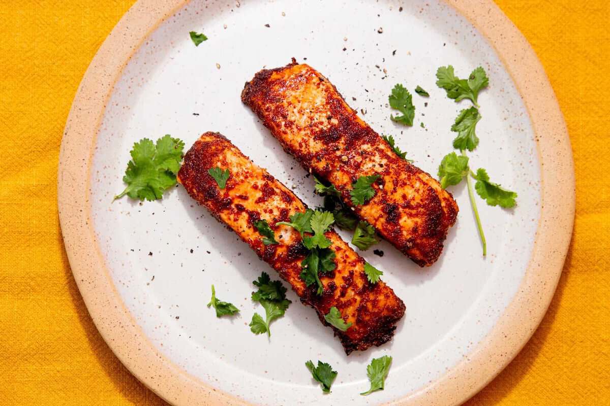 Two pieces of seasoned, grilled salmon garnished with fresh cilantro are served on a white plate with a beige rim, placed on a yellow surface.